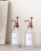 Two glass hand soap dispensers with copper pumps on a neutral background