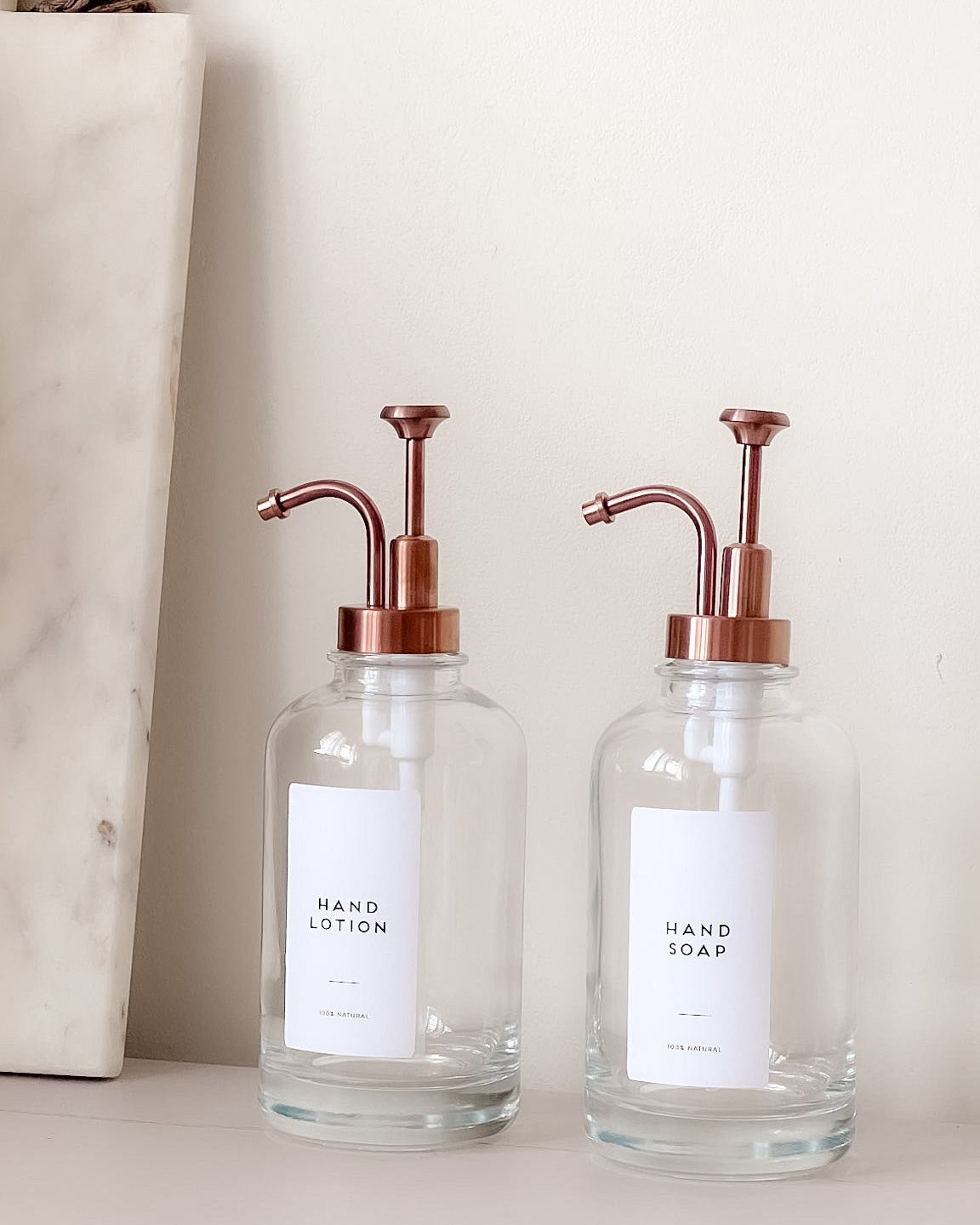 Two glass hand soap dispensers with copper pumps on a neutral background