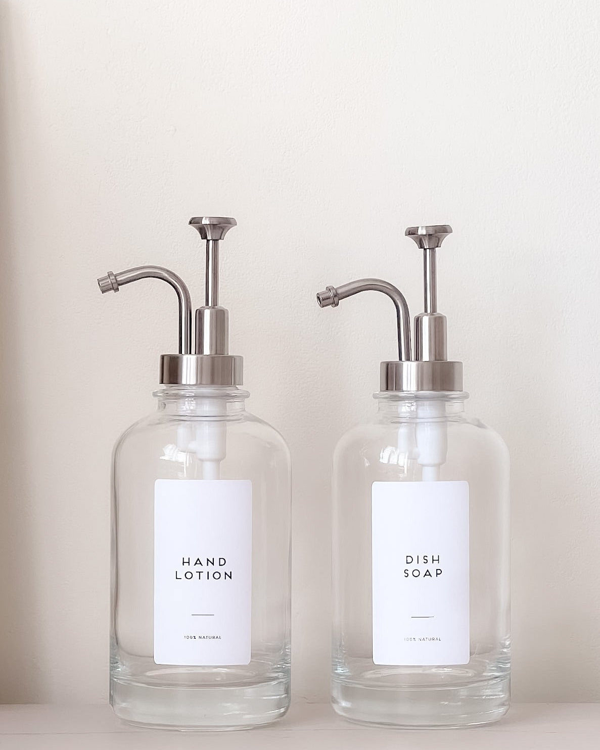 Two glass dispensers with pump tops labeled 'Hand Lotion' and 'Dish Soap' on a neutral background.