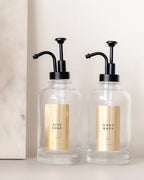 Two glass soap dispensers with black pumps labeled 'Dish Soap' and 'Hand Wash' on a neutral background.